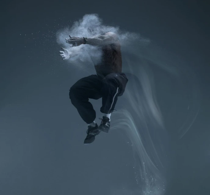 Athletic man mid-air, shirtless in black pants and shoes, surrounded by white powder against a gray background, studio lighting, action shot.
