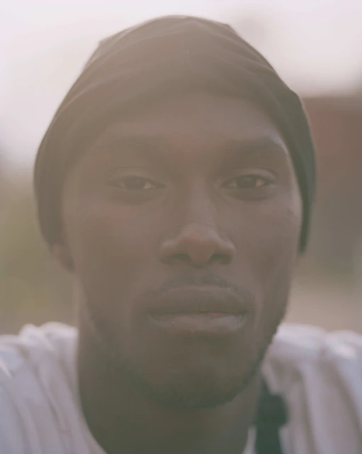 Close-up studio portrait of an adult Black man wearing a black cap, looking directly at the camera with a neutral expression.
