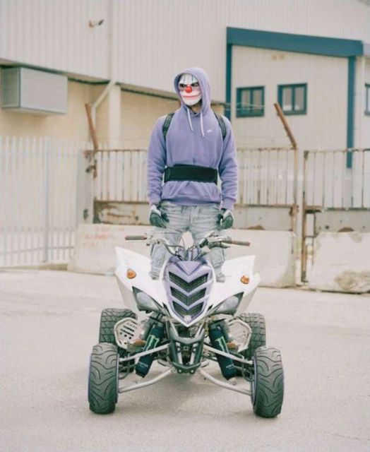 Person in clown mask stands on a white ATV in an urban setting with a white building, gray pavement, and neutral tones.