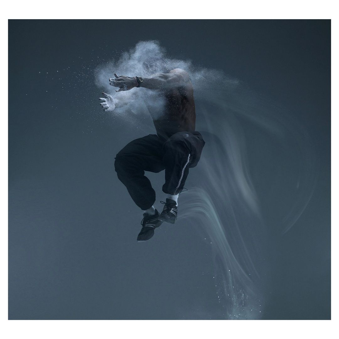 A muscular man mid-air, seemingly suspended in a cloud of white powder against a dark grey background, creating a dynamic, artistic composition.
