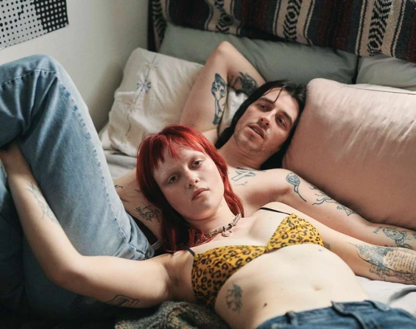 Adult male and female with tattoos pose on a bed in a domestic interior. The woman wears a leopard-print bra. Lifestyle portrait.