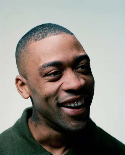 Close-up portrait of a smiling adult Black man with a short haircut, wearing a dark green jacket against an off-white background.