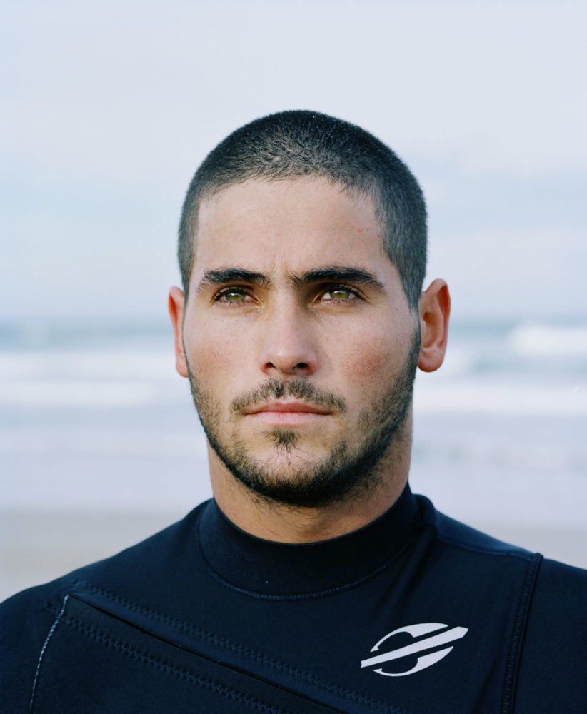 Portrait of a man with dark hair and green eyes wearing a black wetsuit on a beach with the ocean in the background.