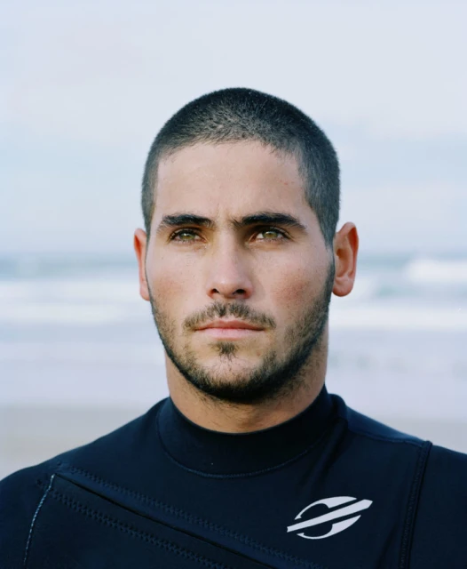 Portrait of a man with dark hair and green eyes wearing a black wetsuit on a beach with the ocean in the background.