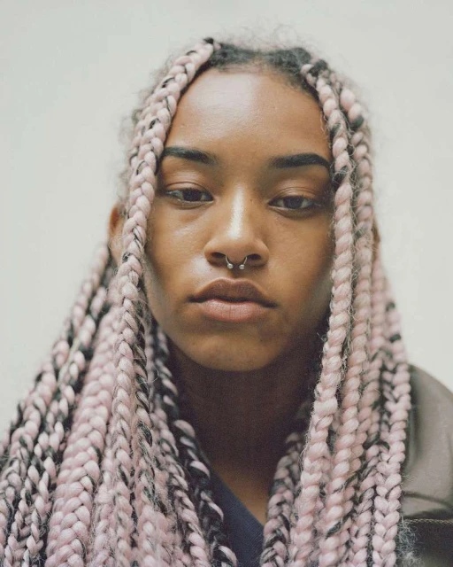 Close-up portrait of a young adult woman with braided hair and a nose ring, posing against a light neutral-colored backdrop in a studio setting.