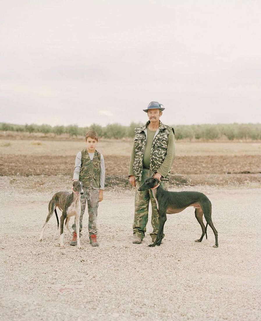 Adult male, child, and two greyhounds stand outdoors. The pair wear camo vests and the dogs have collars, in a rural setting.