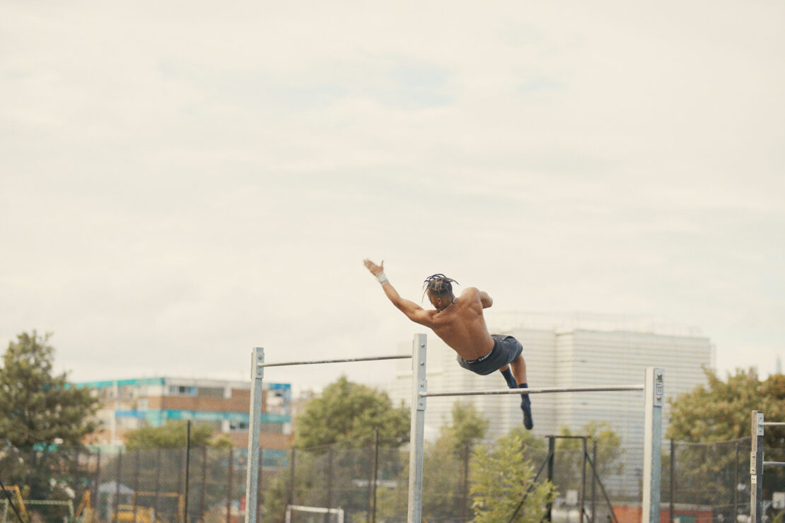 Shirtless man with dreadlocks performs gymnastics on parallel bars outdoors against a backdrop of city buildings and cloudy sky.