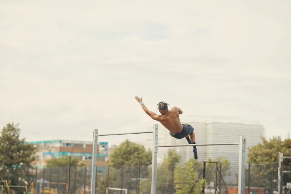 Shirtless man with dreadlocks performs gymnastics on parallel bars outdoors against a backdrop of city buildings and cloudy sky.