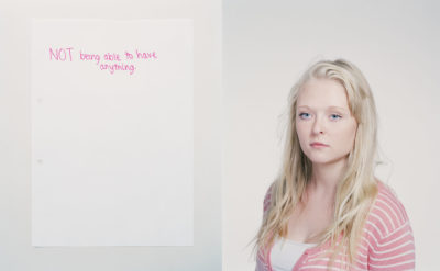 Blonde woman with a serious expression stands next to a piece of paper with handwritten pink text in a bright indoor setting.