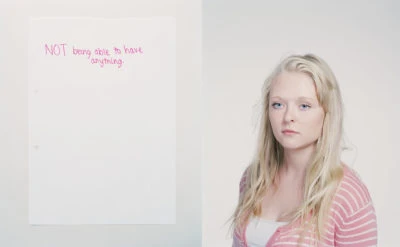 Blonde woman with a serious expression stands next to a piece of paper with handwritten pink text in a bright indoor setting.