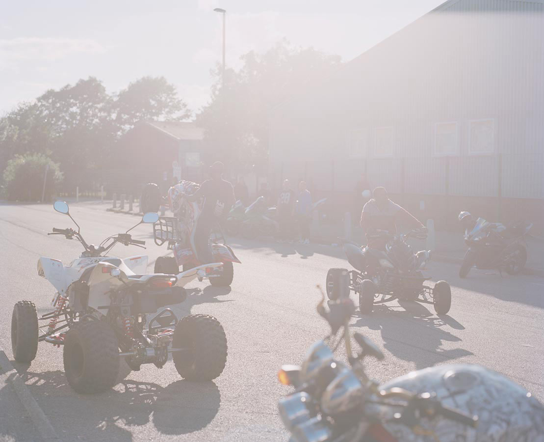 A group of people with ATVs parked on a street in front of a building during daytime with bright sunlight, creating a hazy view.