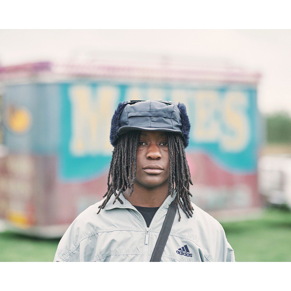 Portrait of a young adult with dreadlocks wearing a fur-lined trapper hat and Adidas jacket, standing outdoors with a blurred fairground background.