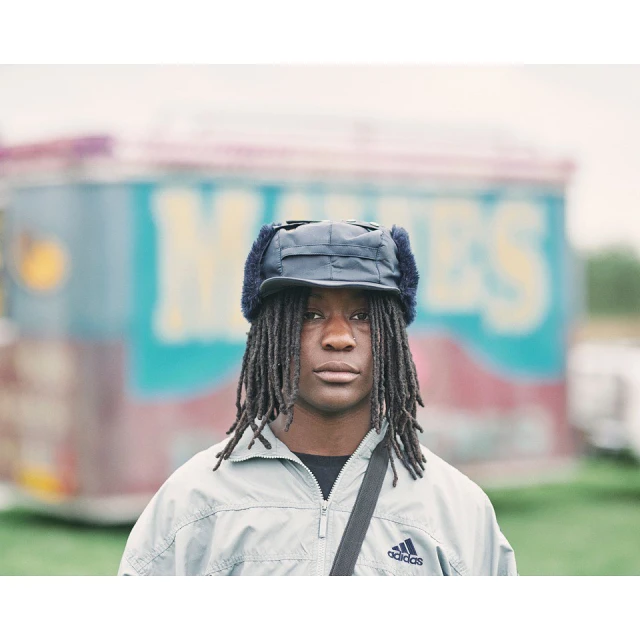 Portrait of a young adult with dreadlocks wearing a fur-lined trapper hat and Adidas jacket, standing outdoors with a blurred fairground background.