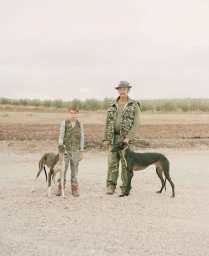 An older man and a young boy standing outdoors in a rural landscape, each holding a greyhound dog on a leash.