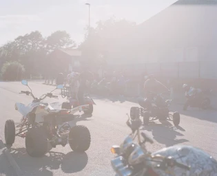 Quad bikes parked on an asphalt street with people standing nearby in bright hazy sunlight by Spencer Murphy.