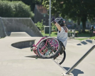 A person in a wheelchair and helmet navigating a concrete skatepark ramp, captured from behind.