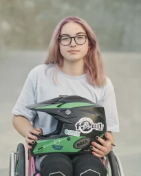 Portrait of young woman with pink hair and glasses holding a black and green motocross helmet while seated in a wheelchair.