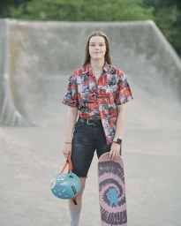 Young woman standing at a concrete skatepark holding a blue helmet and a skateboard by Spencer Murphy in London