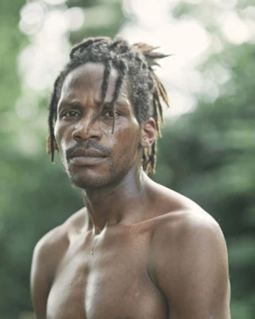 Portrait of a shirtless black man with dreadlocks tied back, captured outdoors against a soft green foliage background.