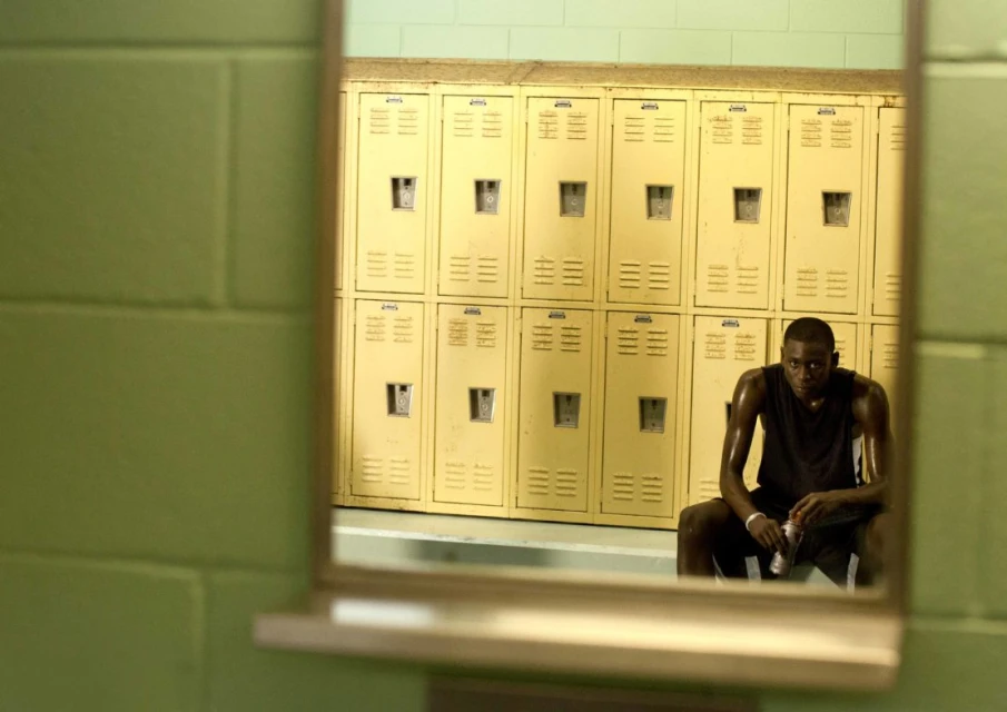 A sweaty man sits on a bench in a locker room reflecting in a mirror, next to yellow lockers and a green cinder block wall.