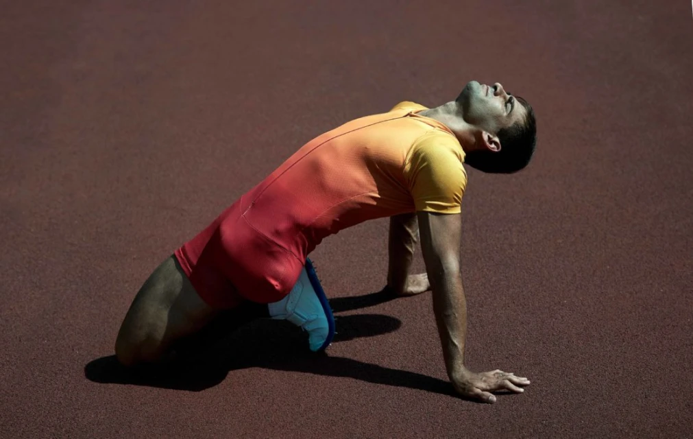 Athlete in a red and yellow uniform kneels on a red track, arching his back and gazing upward in an outdoor setting.