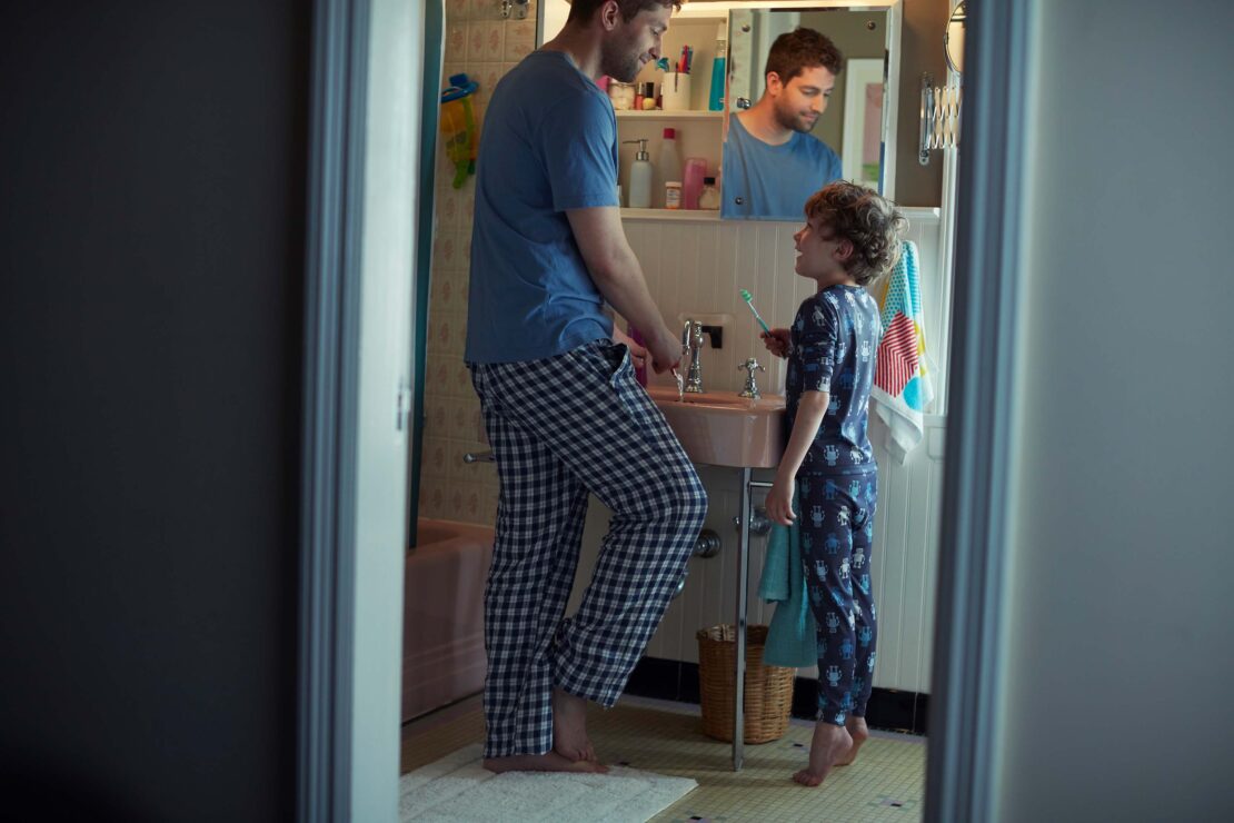 Father and son in matching pajamas in the bathroom, brushing their teeth in front of the mirror in the morning light.
