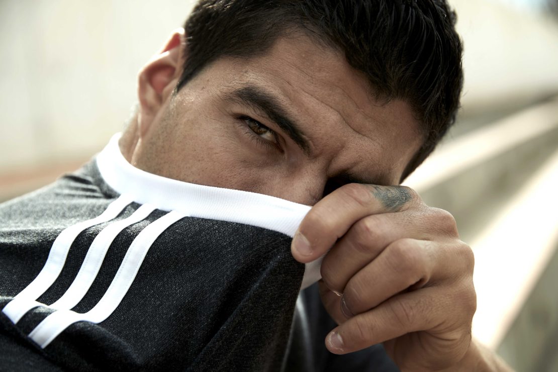 Close-up of an adult male partially concealing his face with a grey and white athletic shirt while staring intently at the camera.