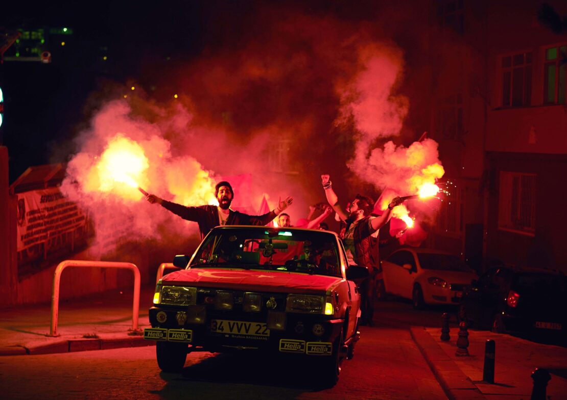 A group of adults around a red car at night, holding burning flares, with red smoke filling the air on an urban street.