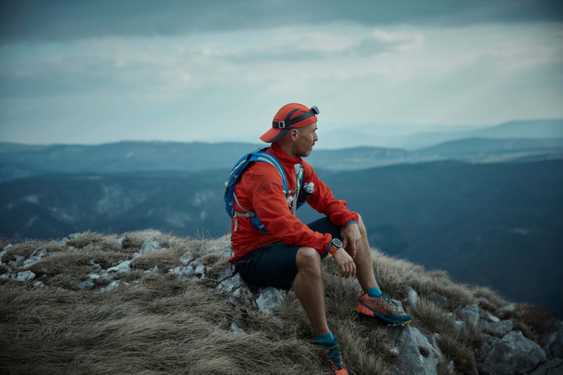 Man in orange jacket sits on a rocky mountain peak overlooking a range of blue hills under an overcast sky, wearing a cap and backpack.