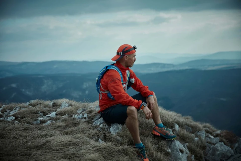 Man in orange jacket sits on a rocky mountain peak overlooking a range of blue hills under an overcast sky, wearing a cap and backpack.