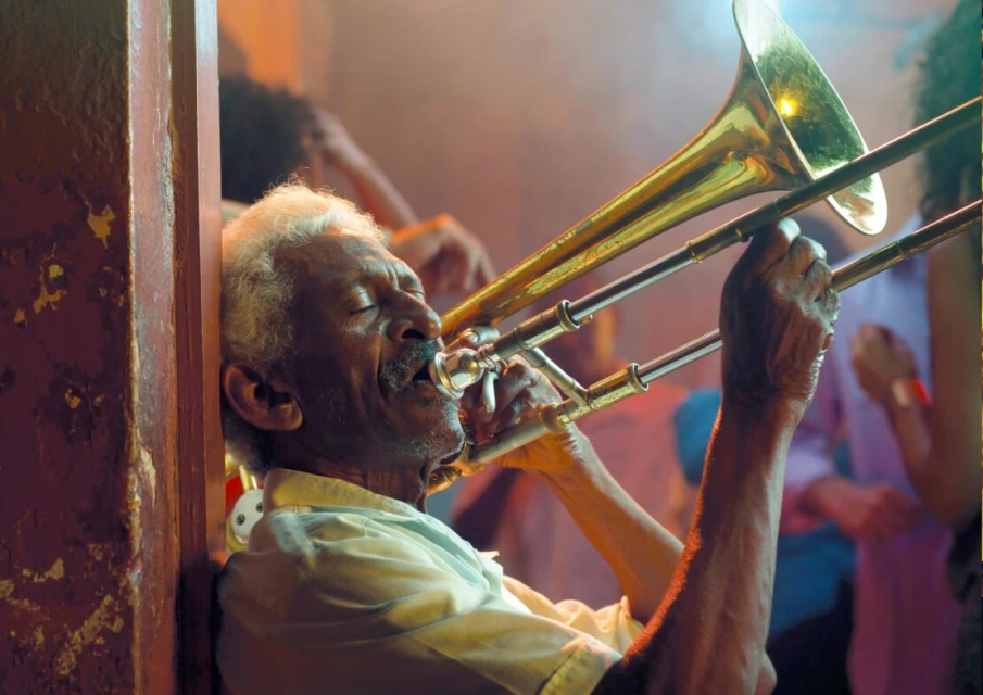 Elderly African-American man plays a shiny brass trombone inside a venue with a blurred crowd and orange lighting, showcasing musical talent.