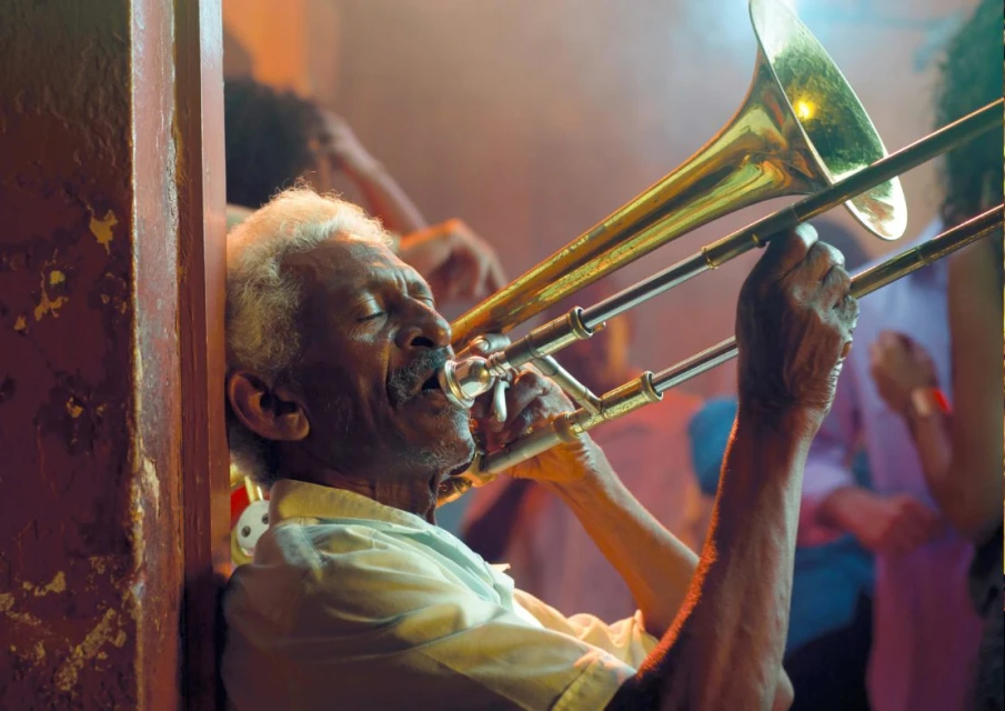 Elderly musician plays trombone in a dimly lit venue, with visible hand textures, brass instrument details, and blurred background figures.