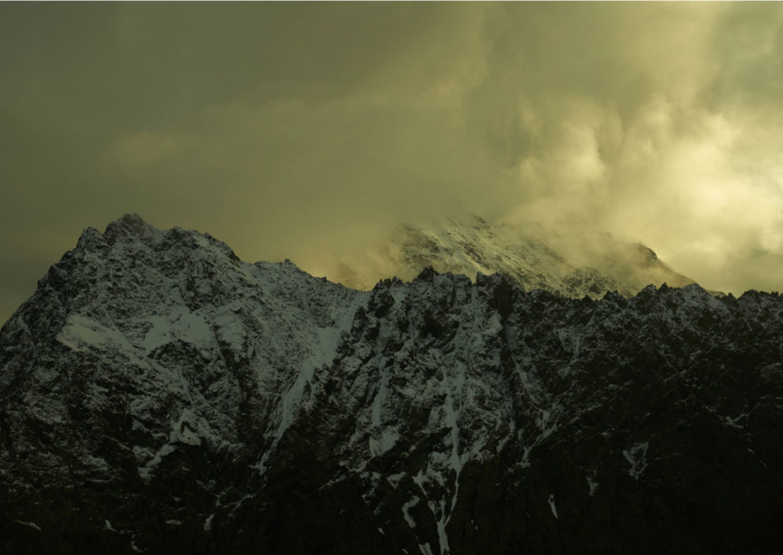 Snow-capped mountain peaks rise under cloudy skies, showcasing a rugged, natural landscape with dramatic atmospheric conditions.