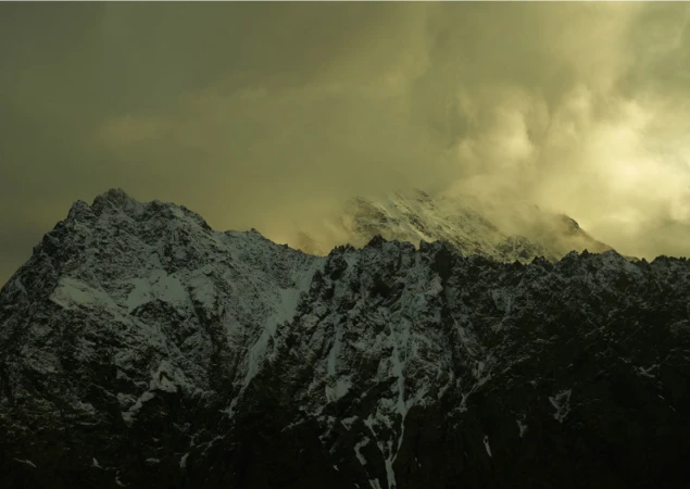 Snow-capped mountain peaks rise under cloudy skies, showcasing a rugged, natural landscape with dramatic atmospheric conditions.