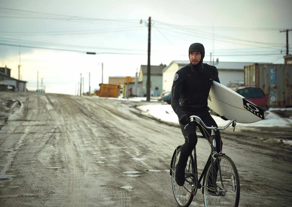 An adult male in a wetsuit rides a bicycle carrying a surfboard through a snowy village street on a cloudy day near buildings.