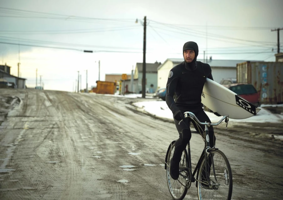 An adult male in a wetsuit rides a bicycle carrying a surfboard through a snowy village street on a cloudy day near buildings.