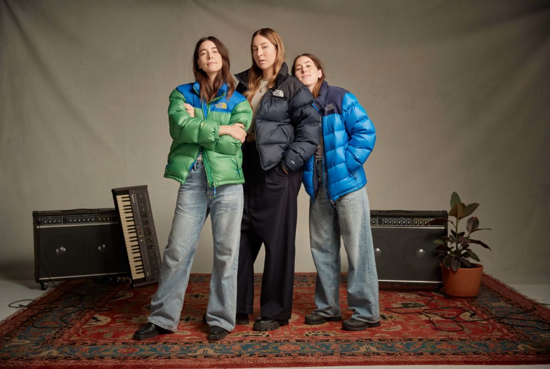 Three women pose in a studio wearing puffy jackets and jeans, standing in front of musical equipment on a red patterned rug.