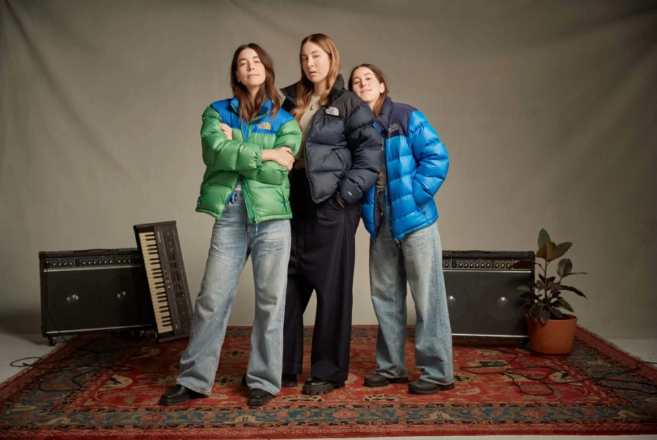 Three women pose in a studio wearing puffy jackets and jeans, standing in front of musical equipment on a red patterned rug.