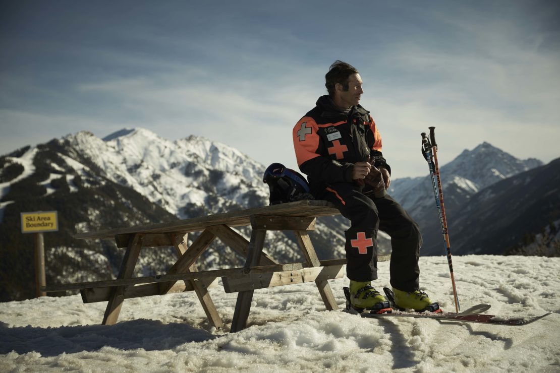 Adult male skier in ski resort uniform sitting on a wooden picnic table with ski equipment; snow-covered mountain background and blue sky.