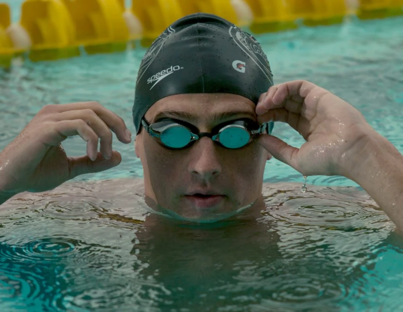 A swimmer in a pool wearing a black Speedo cap and goggles, adjusting goggles with wet hands in the water, looking straight ahead.
