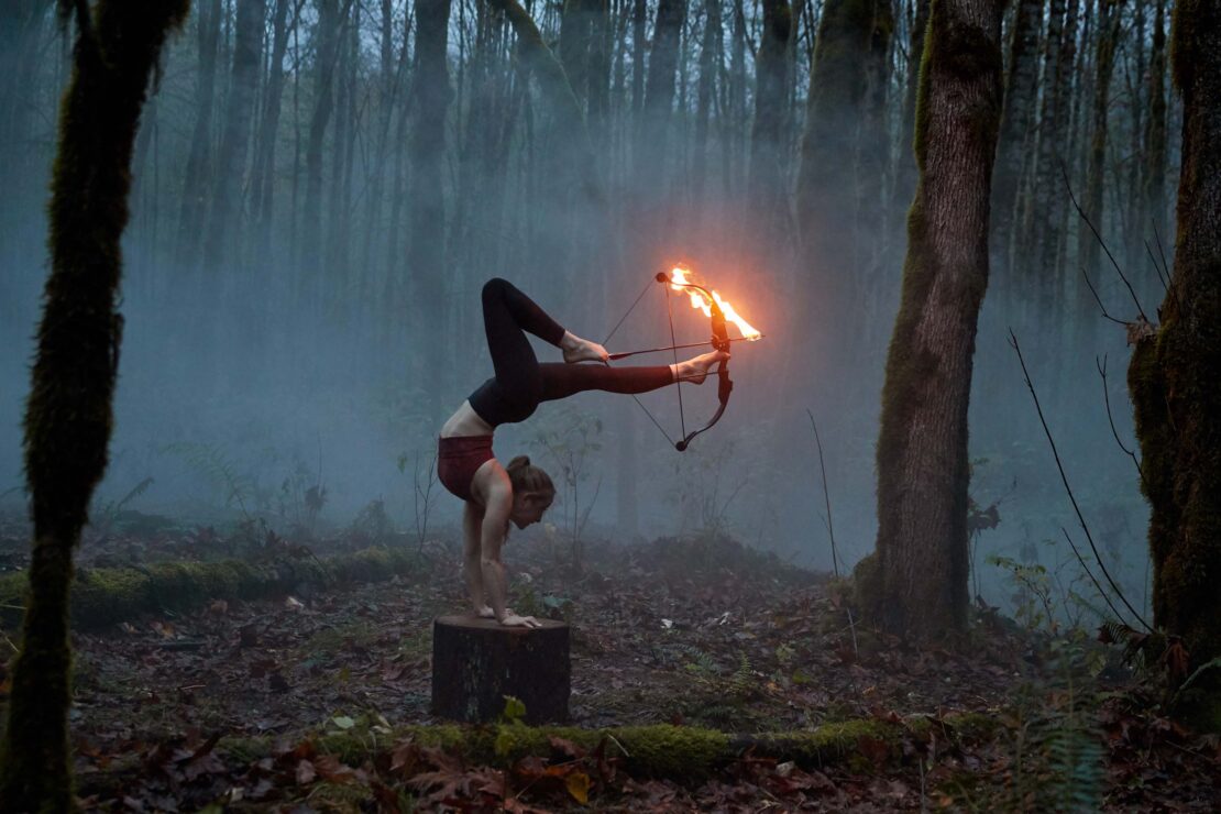 A flexible woman performs a handstand on a tree stump while holding a fire-lit bow in a misty forest setting at dusk.