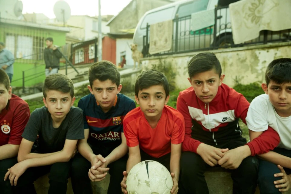 Group of young male soccer players sitting outdoors in a residential area with a soccer ball, captured in a medium shot with natural lighting.