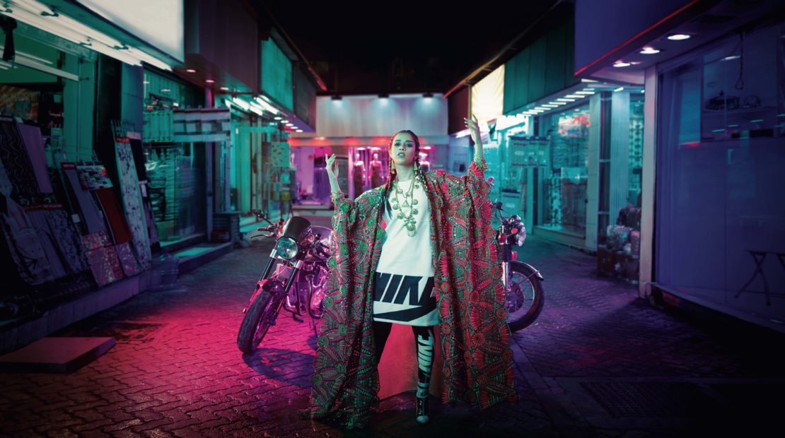 Fashionable adult female in an urban alleyway, wearing a printed robe and jewelry, posing near two motorcycles under neon lighting.