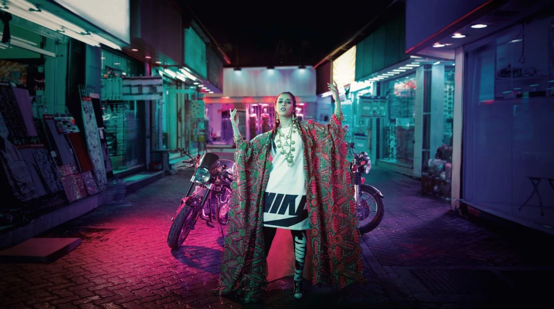 Fashionable adult female in an urban alleyway, wearing a printed robe and jewelry, posing near two motorcycles under neon lighting.