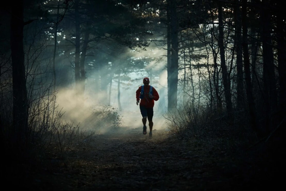 Man trail running through a misty forest. Adult male in red jacket and shorts with headlamp. Forest with tall trees and low-lying ground fog.