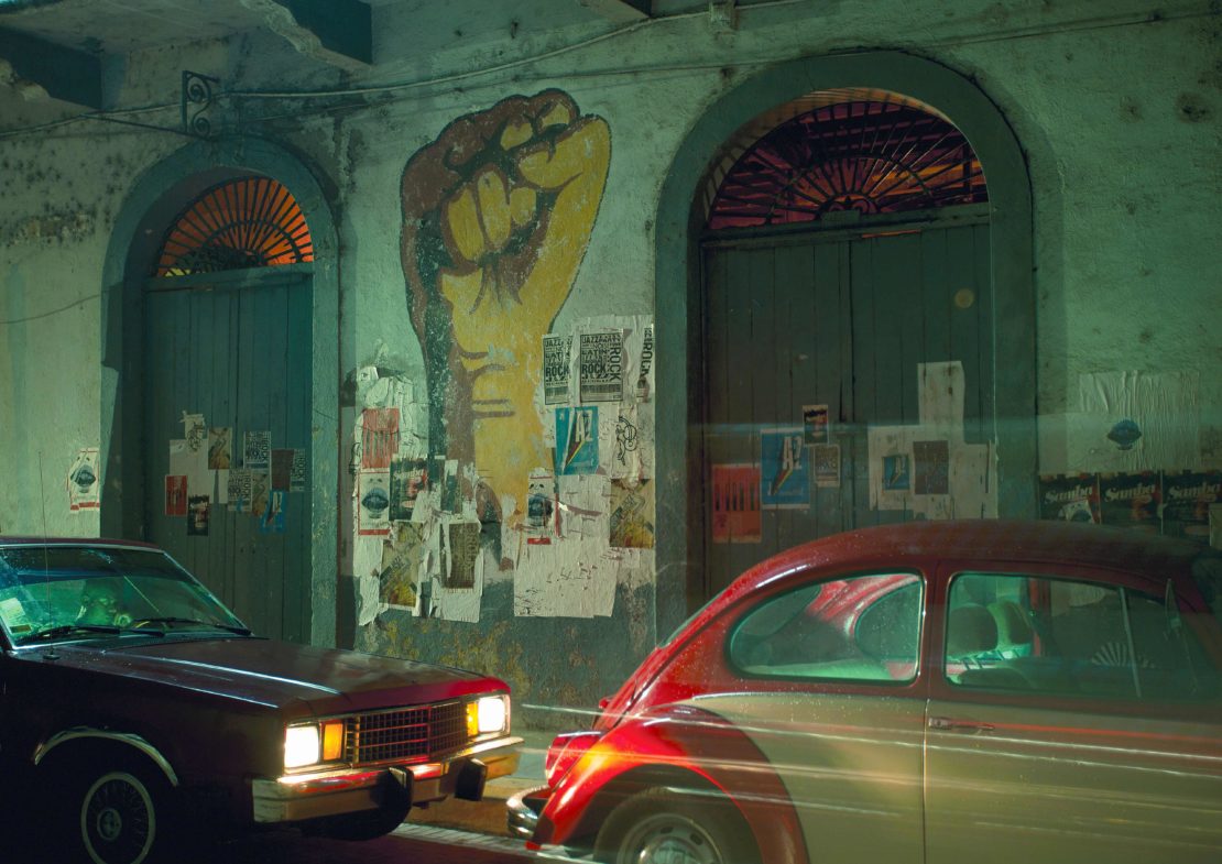 Urban scene at night features a hand mural on a building flanked by vintage cars and posters, illuminated by street lighting.