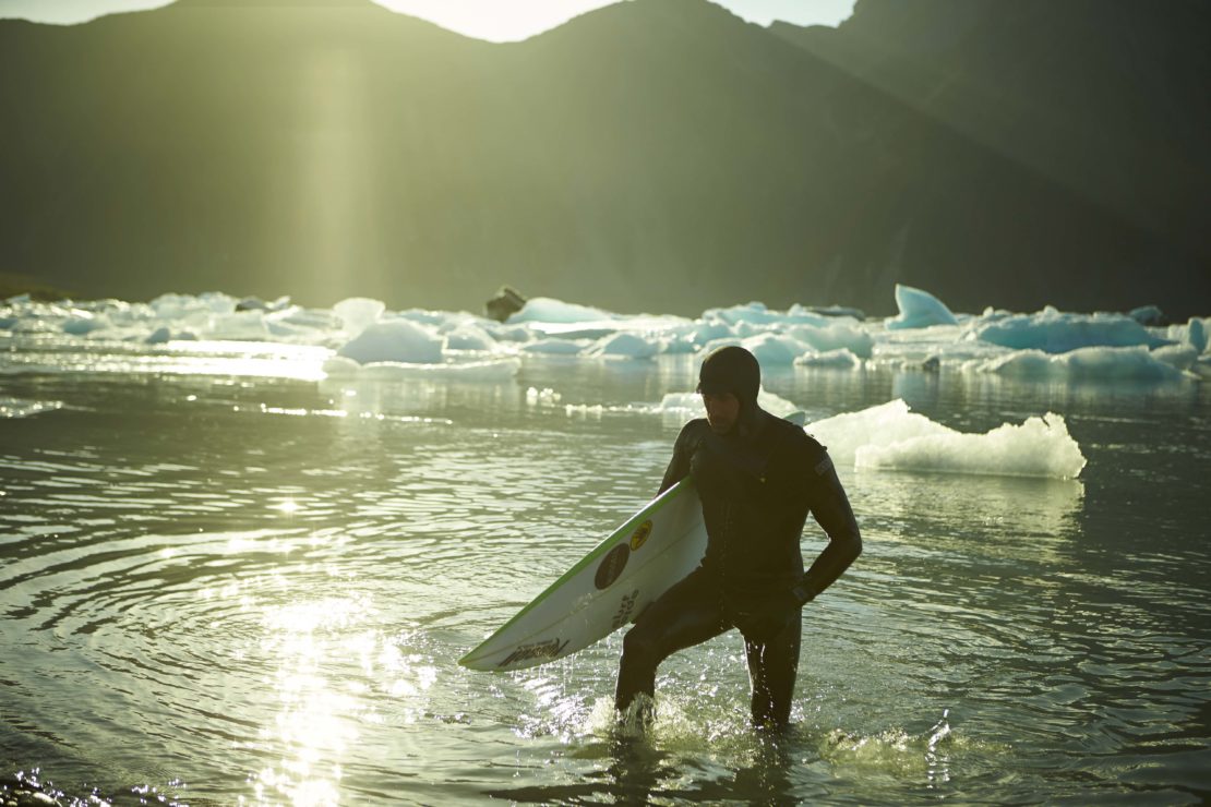 Surfer in a black wetsuit walks out of water towards the shore, carrying a surfboard, with mountains and icebergs in background.