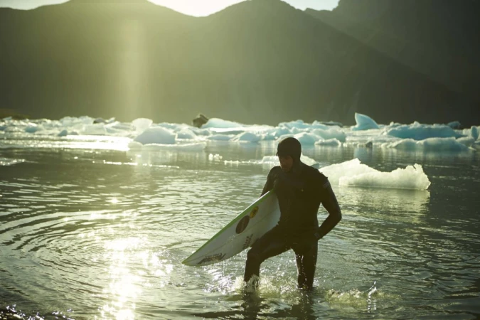 Surfer in a black wetsuit walks out of water towards the shore, carrying a surfboard, with mountains and icebergs in background.