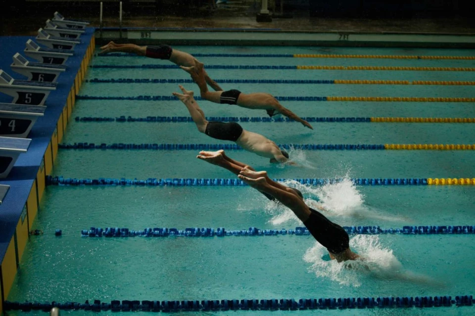Three male swimmers dive into an indoor swimming pool for a race, creating splashes and ripples in the water, with starting blocks visible.