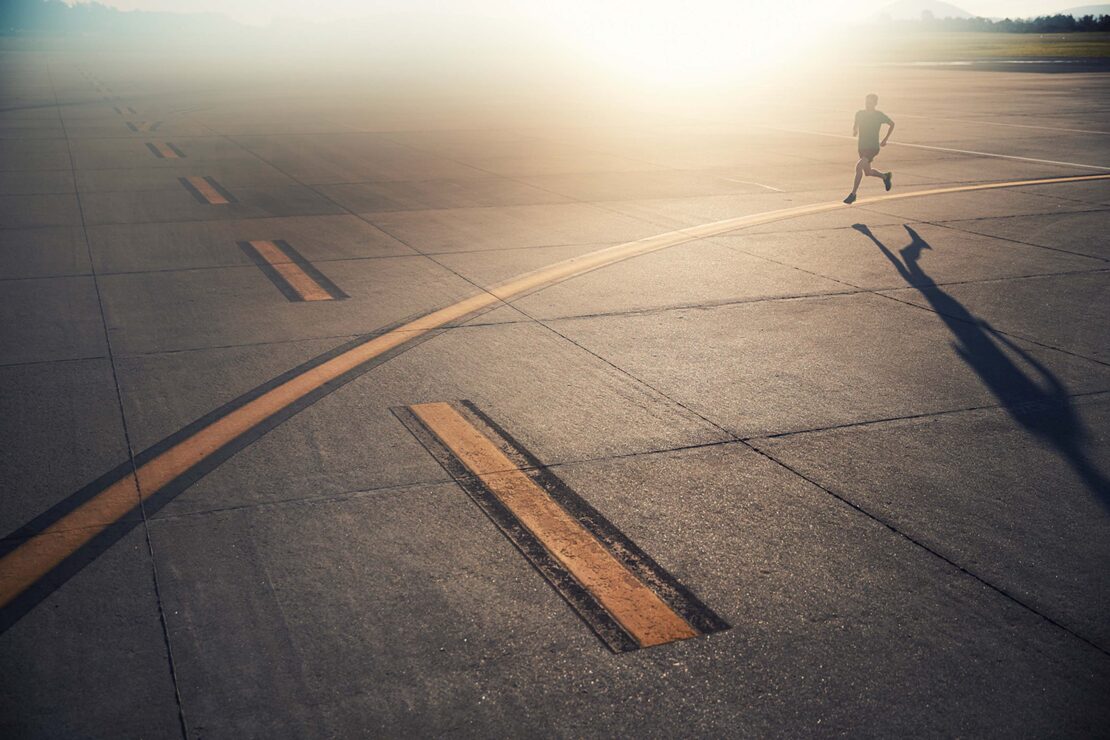 Adult male running outdoors on concrete surface with yellow markings in bright sunlight. Shadow visible. Distant mountains.